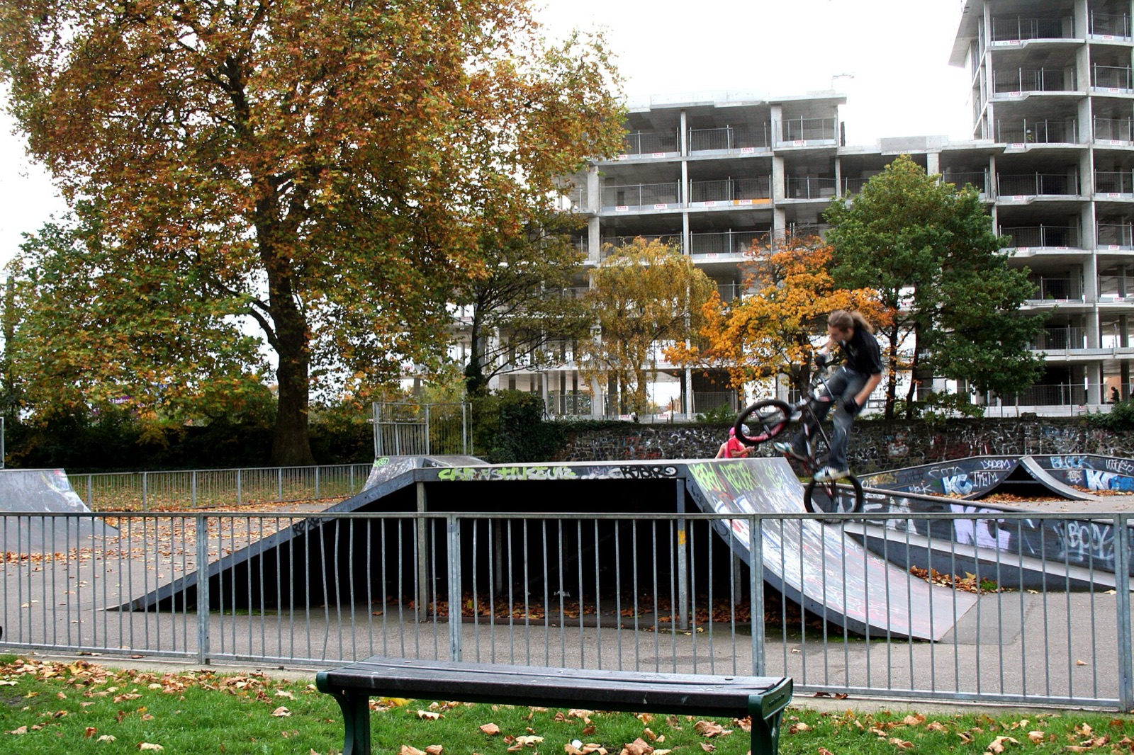 A BMX rider jumps off a ramp at a fenced park skatepark surrounded by autumn trees and flats.