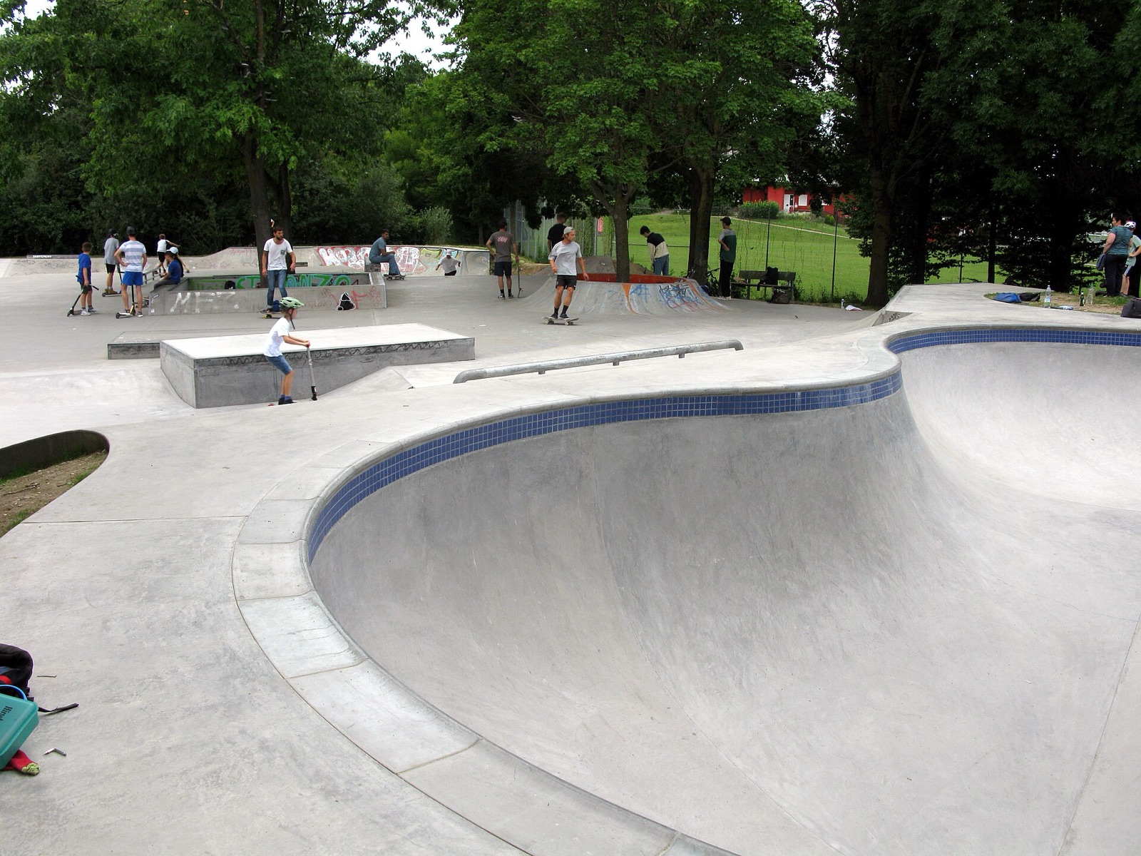 A large tiled-edge concrete skate bowl in a public park, with young skaters and scooter riders on the flat area beyond.
