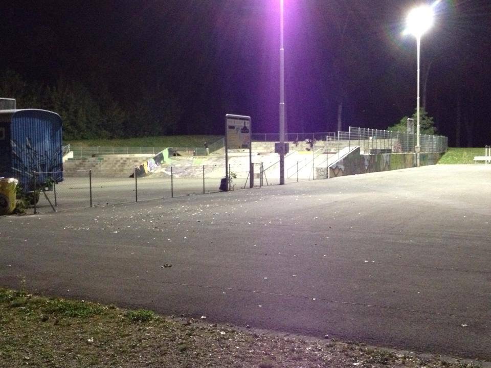 A skatepark lit by tall floodlights at night, with ramps and railings visible across the concrete surface.