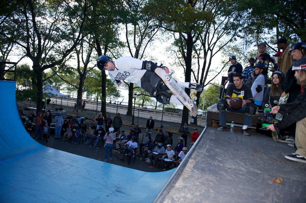 A skateboarder grabs air above a blue mini ramp while a crowd of spectators sits and stands along the side watching.