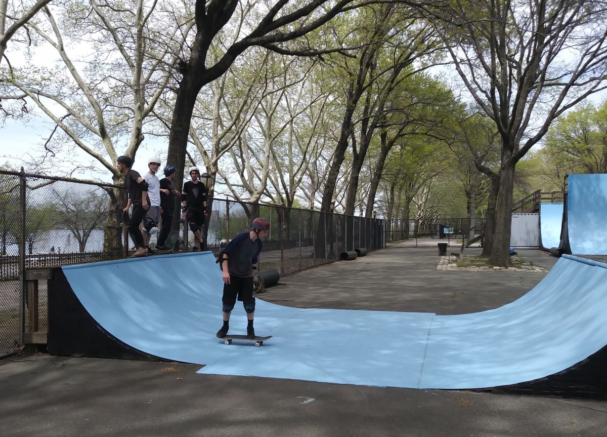 A skateboarder rolls across a pale blue mini ramp under trees while other skaters wait on the deck.