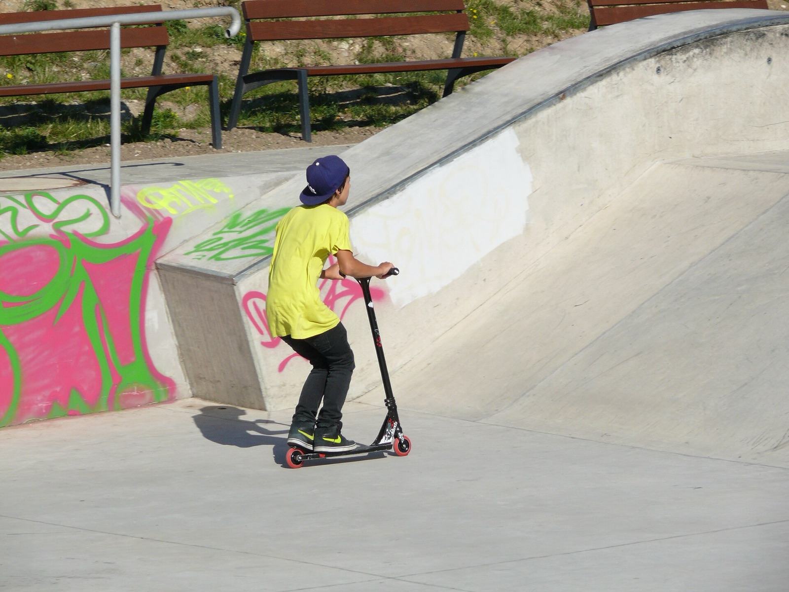 A boy in a yellow t-shirt and cap rides a scooter towards a concrete quarter pipe with benches above.
