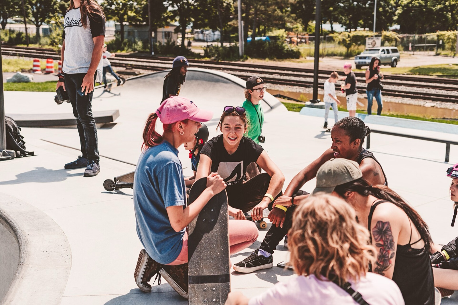 A group of young women and girls sit and chat together on the concrete at a busy outdoor skatepark.