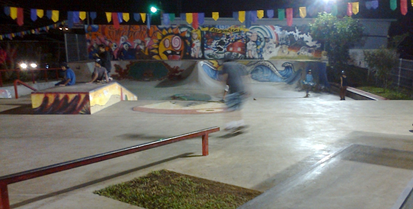 A skatepark at night under lights and bunting, with a low grind rail in the foreground and skaters by painted ramps behind.