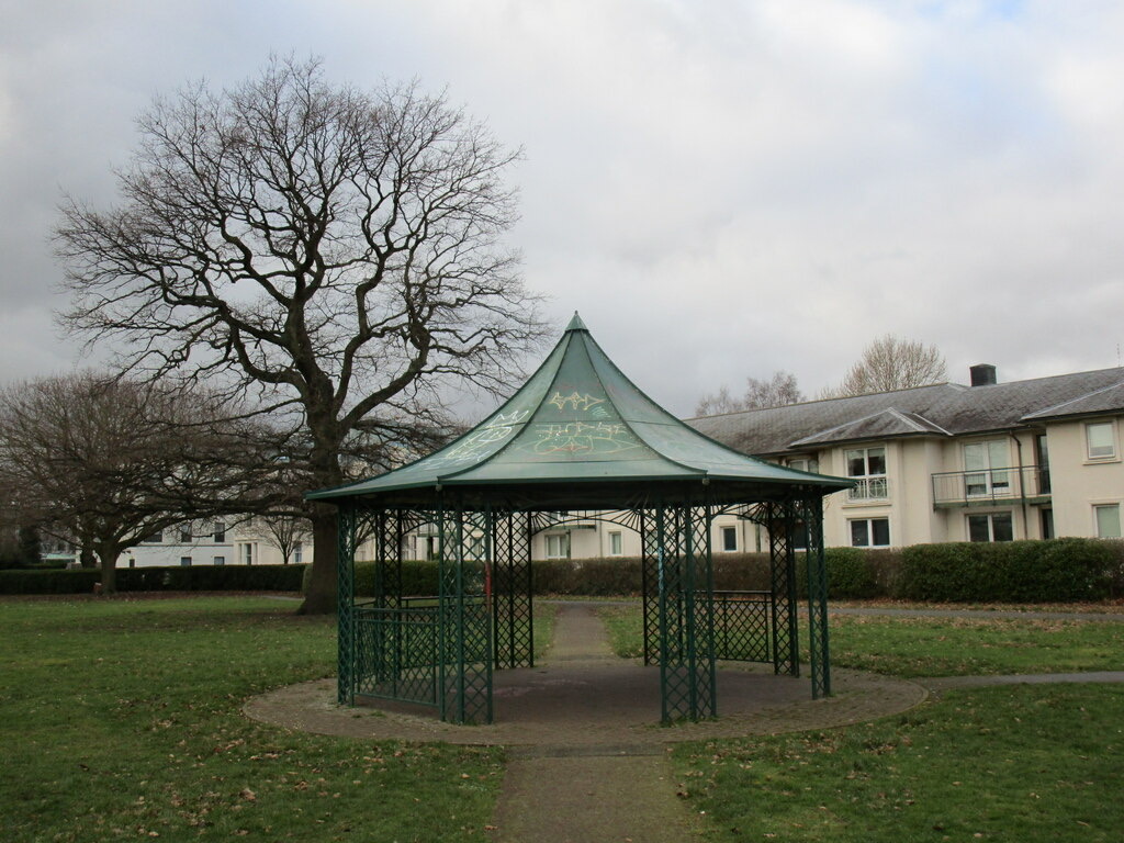 A green metal gazebo on a paved circle in Southfields Park, Loughborough, under a large bare tree on an overcast day.