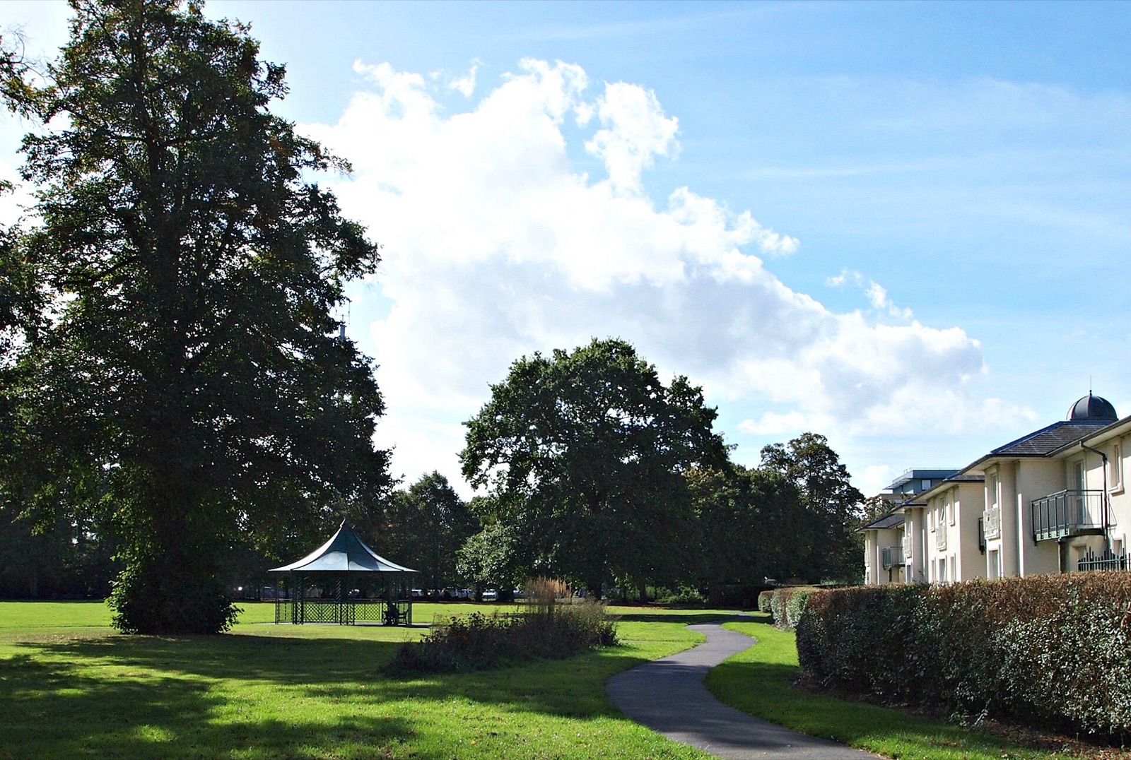 Open lawns and mature trees in Southfields Park, Loughborough, with a path leading to the green gazebo and flats on the right.