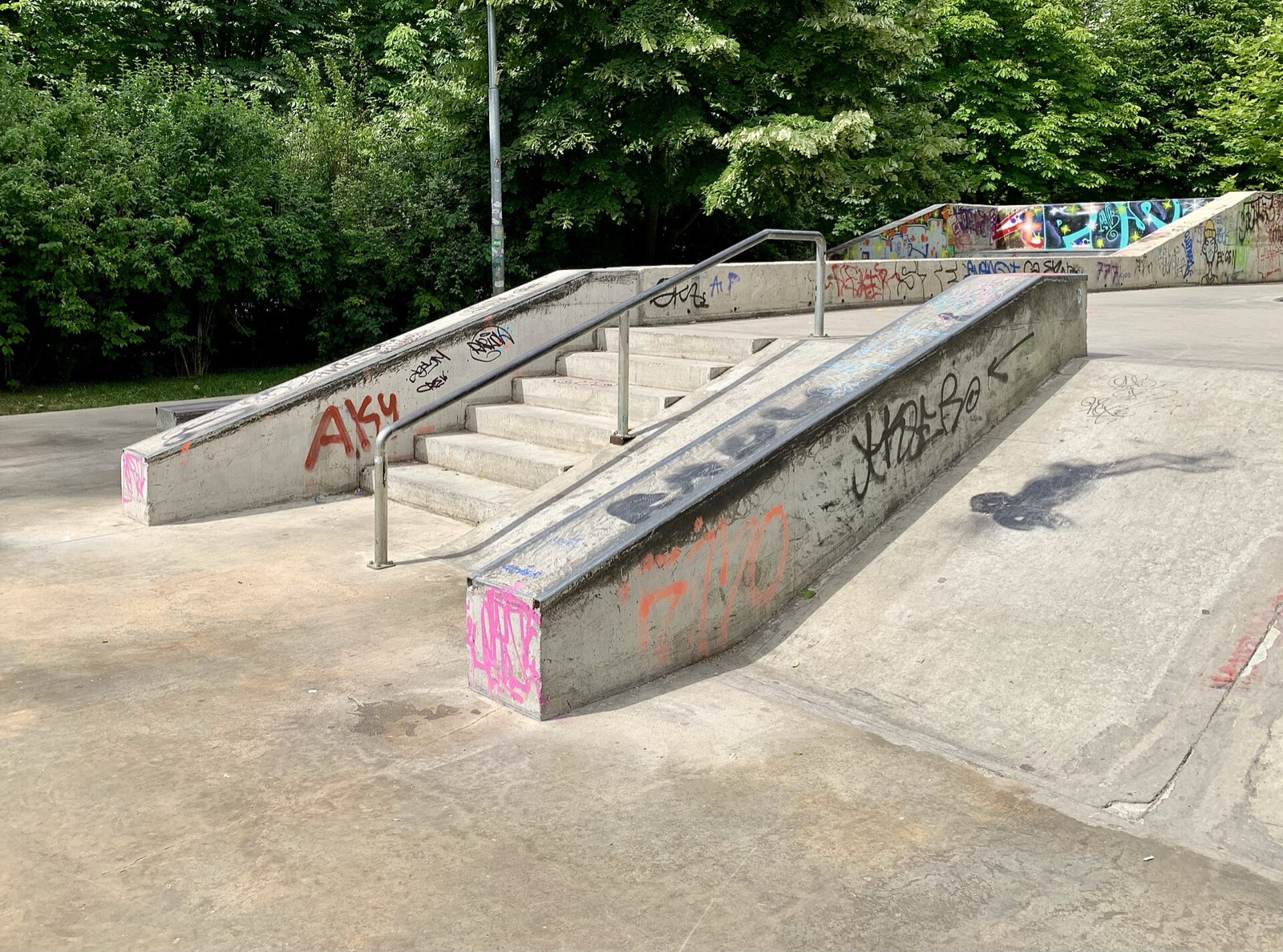 A concrete street section at a public skatepark with a set of stairs, a handrail, ledges and a bank.