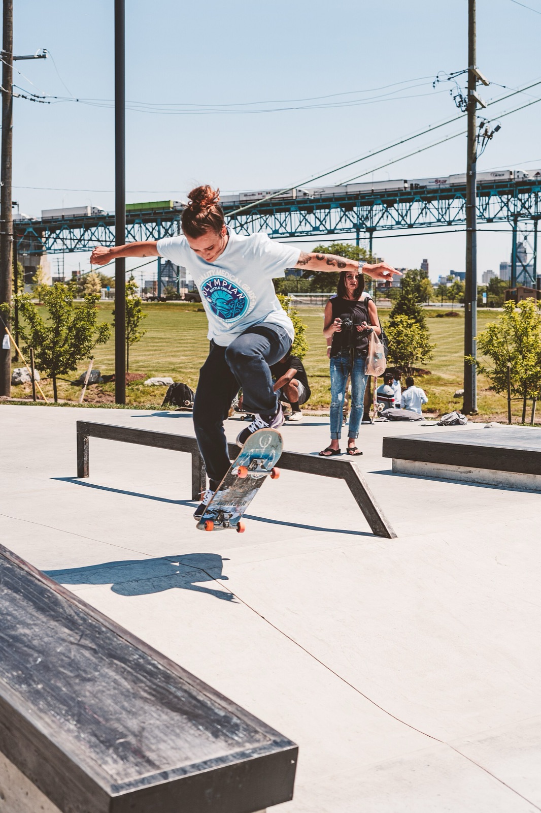 A woman skateboards along a low ledge at an outdoor concrete skatepark on a sunny day, with a bridge in the distance.