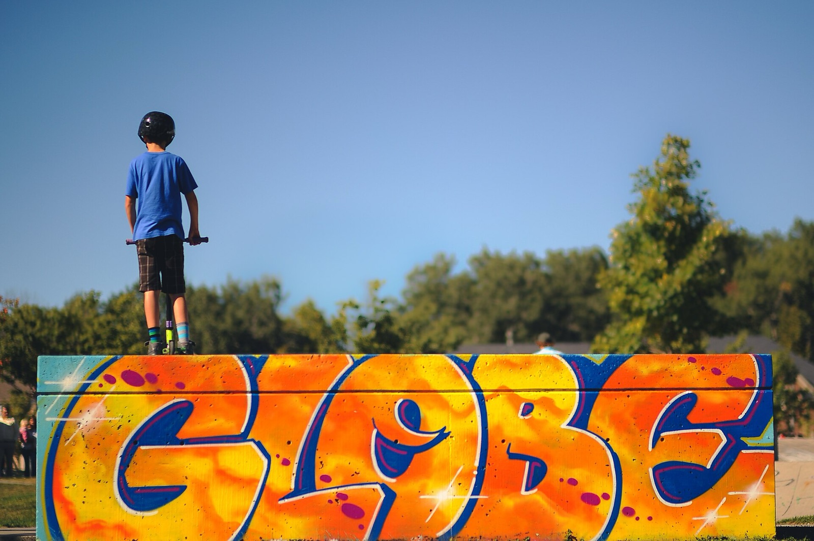 A young child in a helmet stands with a scooter on top of a graffiti-painted box at a skatepark, seen from behind.